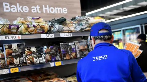 EPA A Tesco employees stands with their back to a set of shelves showing different types of bread for sale in the shop. The employee is wearing a blue hat and fleece, both of which have Tesco branding.