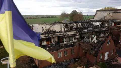 Getty Images A Ukrainian flag flies from a building opposite a hotel used to house refugees from Ukraine that is now a charred ruin following what police suspect was an arson attack days before in the hamlet of Gross Stroemkendorf on October 24, 2022 n