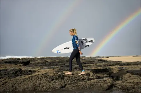 Alan van Gysen/Getty Images Stephanie Gilmore walking with her surf board near a rainbow.