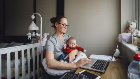 Getty Images Woman working from home at a desk in her bedroom. She is using a laptop while holding her baby daughter
