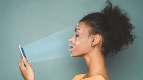 Getty Images A woman holds up her phone in front of her as it scans her face with points of light in this photo illustration combination