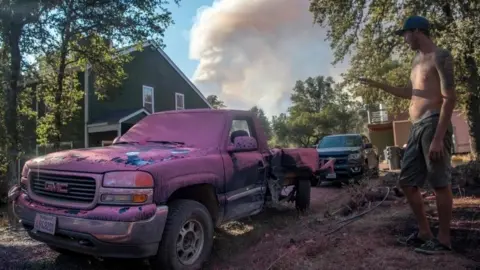 AFP A resident looks at his truck covered in fire retardant as a smoke plume billows in the background near Oroville, California (08 July 2017)