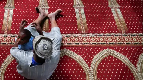Reuters An overhead shot of a boy looking at the camera while he sits with his father inside a mosque.