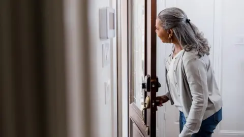 An older woman is opening a door and looking out at someone from behind it. She is wearing a white cardigan over a cream top and has grey shoulder-length hair.