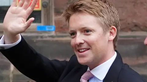 PA Media Hugh Grosvenor, who has ginger spiky hair and is wearing a dark suit jacket over a white shirt, smiles at someone out of frame and waves his right hand