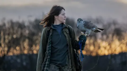 Roadside Attractions A still from the H is for Hawk film. It shows Claire Foy standing in a field while holding a hawk. She is looking away from the camera into the distance. 