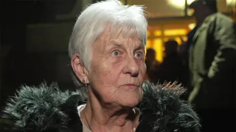 Barbara Crilly standing outside the church at night. She has short white hair and is wearing a black coat with fur-lined hood