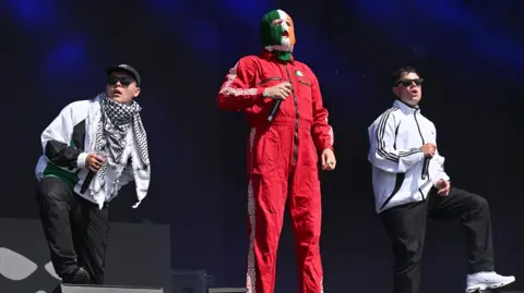 Leon Neal/Getty Images Mo Chara, DJ Próvaí and Móglaí Bap on stage at Glastonbury in June. Mo Chara and Móglaí Bap are dressed in white sports jackets and black trousers and are each resting a foot on a speaker. DJ Próvaí stands between them dressed in a red jumpsuit and a striped green, white and orange balaclava.