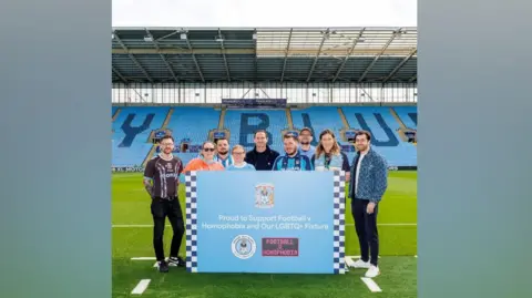 Proud Sky Blues Members of the Proud Sky Blues group with Frank Lampard standing in the middle. A banner is held in the middle with the Coventry City emblem at the top and the text reading 'Proud to Support Football v Homophobia and our LGBTQ+ Fixture'.