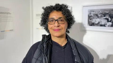 Samantha Walker is a middle aged lady. She has dark skin with curly black hair and wears black glasses. She is standing in front of a photography exhibition. 