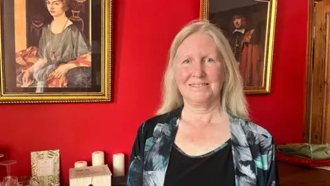 A woman with blonde hair stands in a room with red walls and prints of old paintings on the wall. She is wearing a black top and matching jacket with a floral print design.
