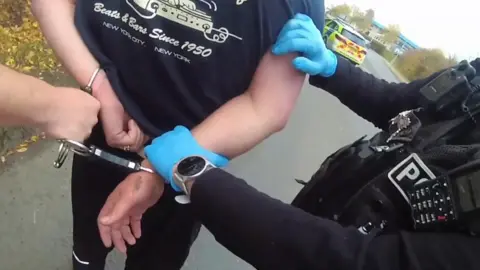 A still image from a police body camera, which shows a man's back, with his wrists being handcuffed behind his back. Someone in black police uniform is holding his wrist with a blue latex glove. The officer is wearing a silver watch. They are outside, and a marked police car is parked in the background.