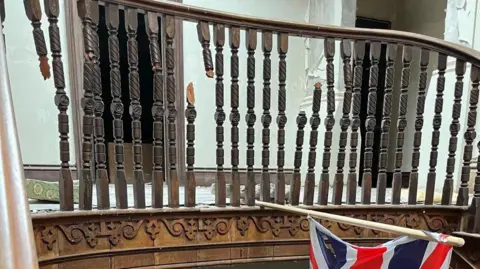 The Somme Association A broken brown ornate staircase with a union flag hanging. The walls are cream and there the ground is rubbly and white. 