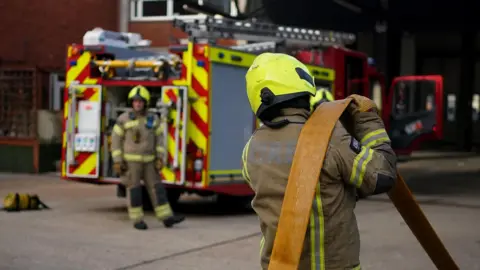 PA Media A firefighter carries a hose over his shoulder as he walks towards a fire engine. Over his shoulder we see another firefighter, facing the camera, walking past the appliance.