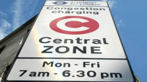A white street sign shown from the bottom looking up. The writing reads 'Transport for London' at the top with a blue Transport for London roundel. Beneath this it reads 'Congestion charging central zone', with a white letter C in a red circle in the middle. At the bottom there is writing reading 'Mon - Fri, 7 am - 6.30 pm'.
