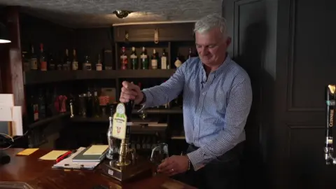 BBC Shot of Richard Knocker pulling a pint behind the bar at the Wolds Inn. The bar area has dark wood units and bottles displayed on a shelf. Mr Knocker has grey hair and is wearing a blue and white gingham-style shirt