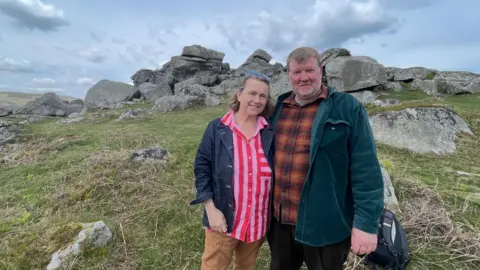Sophie has her sunglasses on her head and wears a bright red and pink striped top. She is next to Alex who is wearing a green jacket. They are both standing in front of Bone Hill Rocks where Alex has just been sketching.