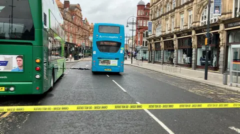 Alex Moss/BBC A road through a city centre with shops on either side. Part of the road has been cordoned off with yellow police tape. In the cordon there are two stationary double decker buses.