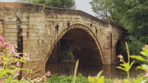 A stone arched bridge, with the River Derwent flowing underneath and pink wild flowers in the foreground, with trees flanking the opposite bank