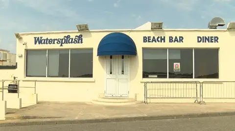 The front of the Watersplash beach bar and diner has those words printed in blue on the yellow building. The white front door has a blue canopy over it. There are three large windows either side of the door and some metal fence panels in front. 