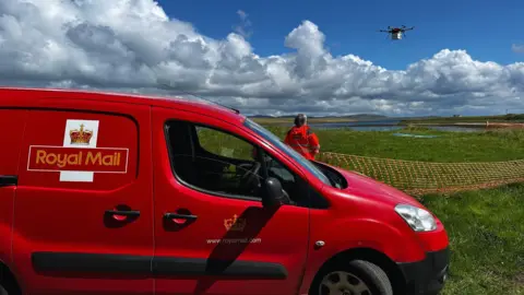 Royal Mail/Press Association An island landscape of flat fields with a sea loch and land in the distance. A Royal Mail van is in the foreground with a person in hi-vis orange jacket behind. The person is looking at a drone in the sky
