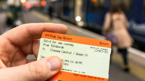 Getty Images A hand holding a train ticket. There are people on the platform in the background which is blurred.