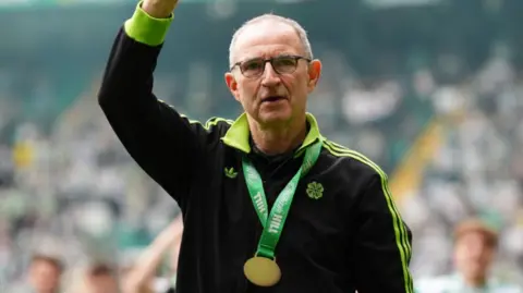 PA Media Martin O'Neill is waving to a crowd inside a football stadium. He is wearing a black tracksuit which has green stripes. Around his neck is a gold medal with a green ribbon. 