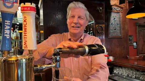 Michael McMahon pulling a pint of stout behind the counter in the Celtic Bar in Washington DC.  He has short, grey hair and is wearing a pink pinstripe shirt. 