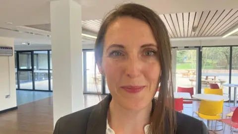 Clare Morris with brown hair wearing a black suit. She is inside a building with yellow and red chairs and white tables.