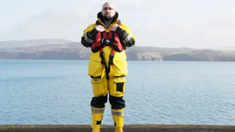 RNLI Cedwyn Rogers stands in front of the water in a yellow RNLI uniform. 