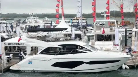 A luxury yacht is pictured docked in a busy harbour. On the jetty a gazebo says 'Sunseeker' on it