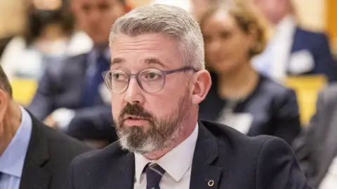 PA Media Niall Ó Donnghaile – a man with gray hair wears a suit, collared shirt, tie and round glasses.