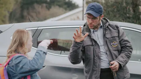 Banjoman Films, supported by Screen Ireland Grainne and Frankie standing next to each other as they share a handshake. Grainne has blonde hair and is wearing a blue denim jacket with a backpack on her back. Frankie is wearing a dark grey jacket, light coloured shirt, black trousers and a blue cap. He is wearing glasses.