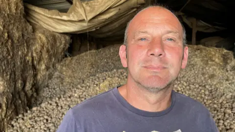 Lynden Croft is wearing a blue T-shirt with 'polo jeans' written on it in a logo. Behind him is a heap of 70 tonnes of potatoes in a big barn.