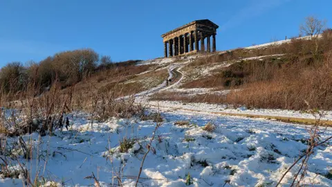 A snow-covered path leads to Penshaw Monument in Sunderland. It is a stone structure with pillars at the top of a hill, resembling a an ancient Greek temple.