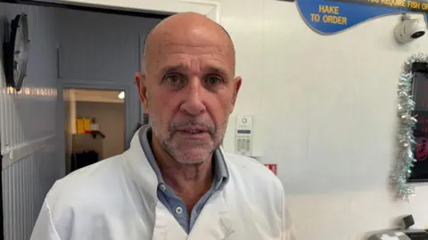 A man with a beard is facing the camera. He's wearing a white chef's coat and a blue shirt underneath. He's standing in his chip shop which has a white tiled wall behind him.