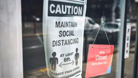 Getty Images A modern sign on a glass door reading “Caution Maintain Social Distancing at least 6ft distance from others” alongside a smaller red sign saying “We’re Closed”.