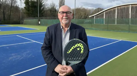 Steve Chilvers standing on a tennis court. He is holding a padel bat. And he is wearing glasses and wearing a suit. 