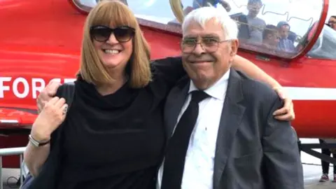 Cull family A picture of Melanie and Raphael Cull, melanie in black dress with sunglasses, raphael in black suit, black tie, white shirt and glasses, both smiling in front a red helicopter