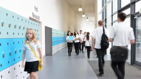 Unhappy schoolgirl walking in school corridor. She wears a yellow cross body bag and has blonde hair. Blurred school pupils can be seen walking away from the camera. Lockers line the hallway. 