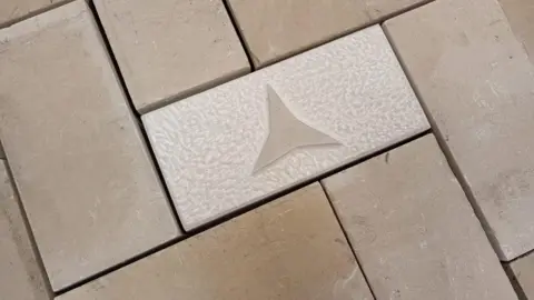 Supplied A white rectangular block, about the same size as a bathroom tile, with a three point star, the symbol of the International Brigade, carved on it. It is surrounded by more blocks.