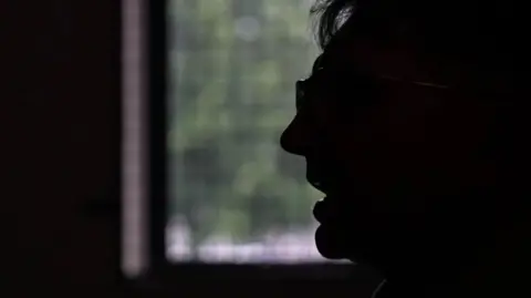 A silhouette of a man's side profile, he has his mouth open as if he is speaking and is wearing glasses. There is a window in the background through which foliage can be seen.