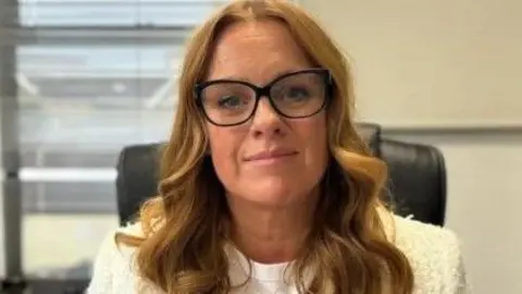 Claire Christian, who has long red hair and is wearing black-rimmed glasses and a white top, is photographed while sitting in an office chair.