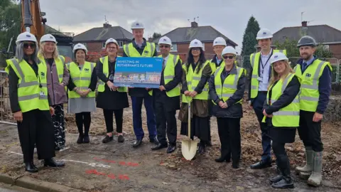 Twelve people, including Council leader Chris Read, wearing high-vis jackets and white hard hats stand on a construction site.