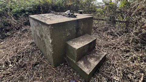David Plaister Ltd A cuboid concrete top of a bunker. It has two concrete steps to the right hand side to allow someone to climb up to the hatch, which is on top and secured with chains. At the far side of this structure is a small metal chimney. The area immediately around the bunker has been cleared recently, as there is bracken on the floor.