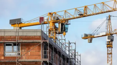 A stock image of a house being built. The house has scaffolding around it and cranes are working on construction.
