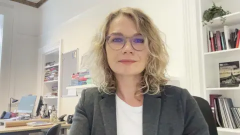 Dr Kelly Johnson, a woman with shoulder-length blonde curly hair and big stylish glasses, looks at the camera while sitting in her office. She wears a white t-shirt and checked wool blazer.