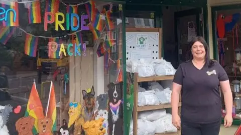 BBC A woman in a black v-neck t-shirt with a bone shaped logo on the upper left side stands in front of a pet shop. To the left the shop window is decorated with pictures of dogs and rainbow lettering that says "Pride pack!". Behind the glass pride flags are visible hanging in the window