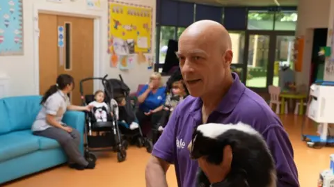 A bald man in a purple shirt holds a black and white skunk in his left arm while smiling. Behind him are several children in prams, with nurses next to them.