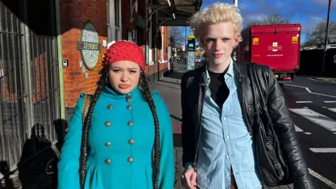 A teenage girl wearing a bright blue coat and red woollen hat, with long hair. She stands next to a blonde-haired teenage boy in a black leather jacket and denim shirt.